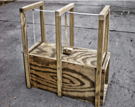 The Armadillo Trap Unscented Wooden Armadillo Trap is designed to capture small animals and features sturdy vertical supports and a string mechanism; it's shown placed outdoors on a concrete surface.
