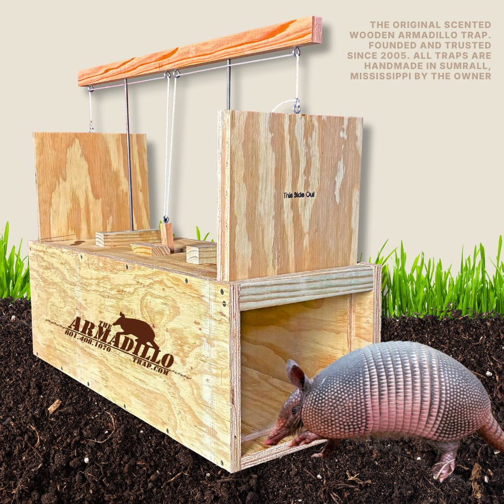 A wooden armadillo trap with open doors sits on soil. An armadillo is near the entrance. Text describes the trap as handmade and scented, made in Mississippi since 2005. The brand logo is on the side.
