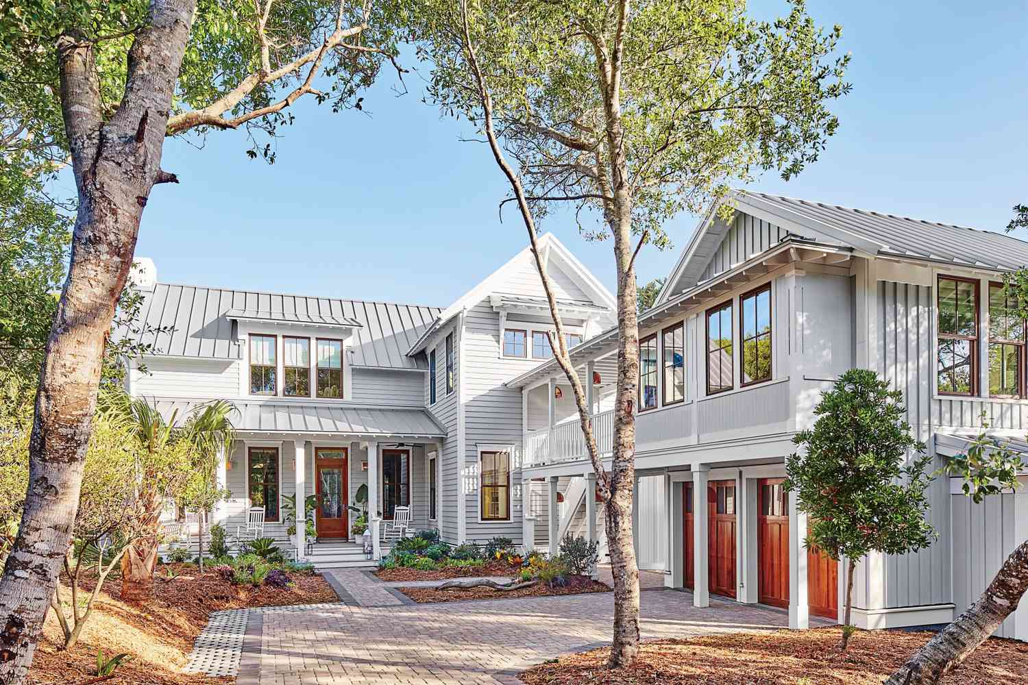 A modern farmhouse-style home with gray siding, white trim, and a metal roof, surrounded by trees and landscaped with mulch and pavers, featuring a front porch and a three-car garage with wooden doors.